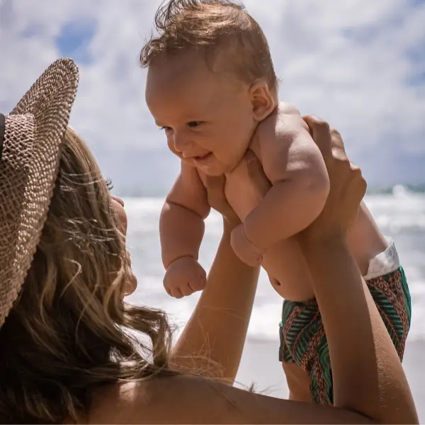 Vrouw tilt lachende baby omhoog op het strand tijdens een zonnige vakantie.